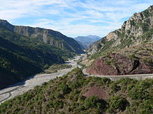 Vue d'une vallée caillouteuse avec une petite rivière, entourée de montagnes rocheuses avec une végétation peu dense.