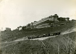 1920 circa - Un convoglio in transito sul tratto Saline-Volterra; sullo sfondo si nota la Fortezza Medicea di Volterra. Più in basso si può scorgere una parte del piazzale della stazione: sulla sinistra sorge la rimessa locomotive; sulla destra sono presenti due carri merci con tetto chiuso e, parzialmente nascosto, il magazzino merci