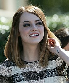 Emma Stone, de face, se passant la main dans les cheveux en souriant.