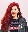 A women with curly red hair stands in front of a pink brick wall.