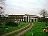 A double track leads through grass towards a small Classical-style building. The central portion has four columns supporting a pediment, and wings with columns extend from each side.