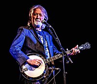 John McEuen, singing into a microphone while playing a banjo.