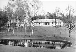 Un bâtiment de plain-pied en fond ; à l'avant-plan, un étang et quelques petits arbres. La photo est en noir et blanc.