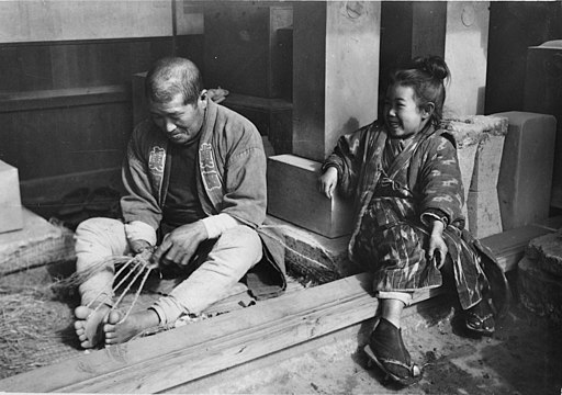 Waraji were traditionally made in the home, as shown here (September 1914). Note twine warp, held between hands and toes, and loose-fiber weft, to his right.