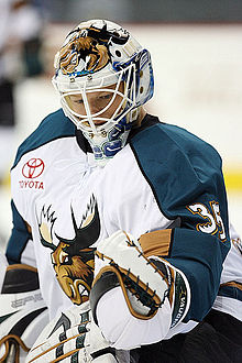 Photographie d’un gardien de hockey, en équipement complet, maillot blanc épaules turquoise, observant son gant