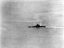 A large area of ocean with a warship in the middle distance. A plume of smoke is coming from the rear of the ship's superstructure, and the ship appears to be leaning to the left.