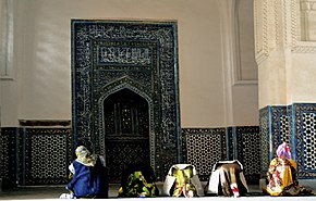 Several women pray inside a building. There is a niche on the wall in whose direction they pray.