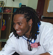 Larry Fitzgerald, un homme noir, avec des tresses, un t-shirt blanc et une boucle d'oreille, sourit à des supporters.