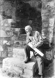 A black-and-white photograph of the architect Bernard Maybeck, dated 1919. In the photograph, Maybeck is gazing at the left side of the frame and resting on a step, slightly leaning to his right with his right foot drawn up on a lower step, the left foot on the ground, and the left hand holding a large rolled paper (possibly a blueprint, indicative of his work as an architect).