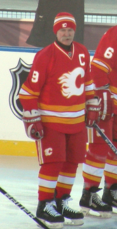 A man stands in a full hockey uniform. It is red with white and yellow trim and a large, white, stylized "C" logo. He is wearing a red toque and has a large moustache.