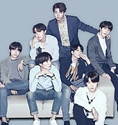 BTS sitting on a white couch with a blue background. The seven members are all wearing various blue and gray outfits.