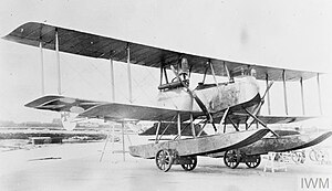 A wooden biplane on the ground, with a small wheeled carriage ("beaching trolley") underneath each float. The Maltese cross is painted on the lower wing and the tail.