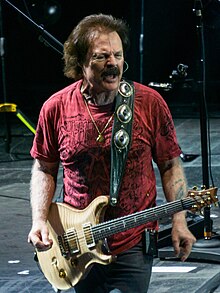 Johnston playing a Paul Reed Smith Artist's Gold Top while wearing a black t-shirt and jeans, in front of colorful stage lights