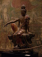 A wooden carving of a sitting Buddhist figure in loose fitting, painted robes.