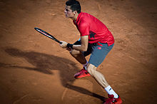 Raonic standing on clay, with his feet wide apart, both hands on his racquet, and looking left. His shadow stretches out sharply to the left.