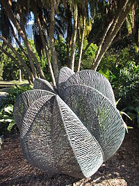 a copper sculpture comprising seven segments wrought out of undulating copper wire, standing beneath a cluster of palm trees