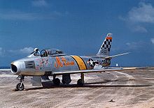 L'avion de chasse argenté de Glenn sur le tarmac, avec une bande jaune derrière le cockpit et un motif en damier sur la queue.