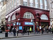 A corner view showing both elevations of a red glazed terracotta building. The first storey above ground features two wide storey height semi-circular windows on each elevation above which is a dentil cornice. Below the windows, the station name, "Oxford Circus", is displayed in gold lettering moulded into the terracotta panels. Above the first two storeys the building has been extended as offices with an ornate Portland stone façade.