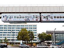 Photo couleur d'un monorail suspendu passant au-dessus de voitures, dans une ville, sous un ciel laiteux.