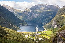 Foto eines Fjords. Der Fjord ist von Bergen umgeben. Im Inneren des Fjords befindet sich ein Kreuzfahrtschiff, am Ufer liegt eine kleinere Ortschaft.