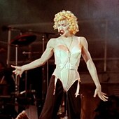 The image of a young blond woman. She is wearing a beige corset and black pants. She has blonde curly hair and has a headset microphone to her mouth.