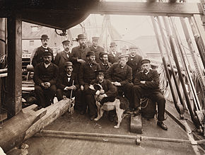 Groupe de 13 hommes et un chien posant sur le pont étroit d'un navire, au milieu des cordes, des épars et du gréement, tous vêtus de chapeau et, à une exception près, de costumes sombres.