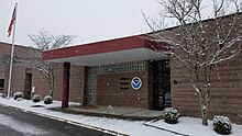 A National Weather Service brick building is seen as snow falls; the ground around the building is already covered with snow, while the road in front of the it is partially covered.