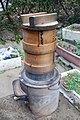 A simple hearth with a metal pan holding two wooden steaming vessels and a wooden lid used in Japan