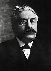 Portrait photographique de trois-quarts d'un homme aux cheveux gris-blancs, aux sourcils et à la moustache broussailleux, portant des lorgnons, un nœud papillon, une chemise blanche, un veston et une veste noire