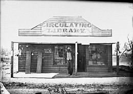 Baraque avec fronton (Circulating Library), homme barbu sur le seuil, pleine campagne.