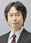 A 67-year-old Japanese man with graying black hair and a suit, looking towards the camera.