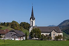 Gambar Gereja Saint Grat yang terletak di Montbovon dengan latar belakang langit biru dan pada bagian bawah gereja tersebut terdapat rerumputan hijau. Di samping kiri dan kanan bawah gereja tersebut, terdapat perumahan dengan atap berwarna coklat dan dinding berwarna putih, dengan dikelilingi oleh pepohonan.