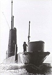 A submarine underway on the surface. A man is standing near the boat's bow sonar dome, while other personnel are visible on top of the sail