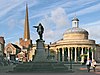 Statue of figure with outstretched arm. To the left a tall church spire and the right a circular building with columns.