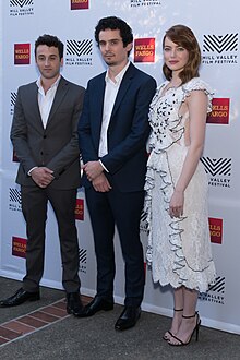 Justin Hurwitz et Damien Chazelle en costume et Emma Stone en robe blanche à pois noirs. Les deux hommes croisent les mains tandis qu'Emma Stone est de profil avec la tête tournée vers nous. Hurwitz regarde le sol tandis que Chazelle et Stone regardent l'objectif.