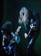 Spotlights pour down on a band, mostly dressed in black, during a performance.