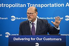 Joe Lhota, ein weißer älterer Mann im dunklem, geschlossenem Sakko, hellem Hemd und Krawatte, steht hinter einem Rednerpult mit dem amerikanischen Slogan „Delivering For Out Customers“ (übersetzt: „Wir liefern für unsere Kundschaft“). Er ist mitten in einer Rede abgebildet, steht vor einem mit Logos der Metropolitan Transportation Authority gefüllten, blauen Hintergrund und gestikuliert mit seiner linken Hand.