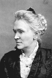 Photographie en noir et blanc, portrait de profil d'une femme blanche aux cheveux blancs en chignon avec un peigne, toilette sombre sur chemisier à dentelle blanc