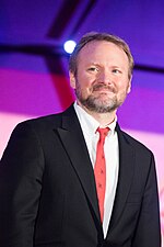 A middle-aged White man with a stubbly beard and mustache; and wearing a dark gray jacket, a red tie, and a white collared shirt; stands in front of a blue, lilac, and white background.