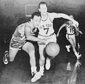 A basketball player chases after a basketball across the court. He stands in front of two other players with jerseys with the words "NEW YORK"