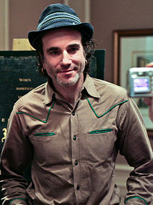 A smiling man wearing a grey hat with piping above the band, and a tan Western style shirt, stands in an office, posing for the camera.