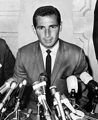 "A man with black hair and in a suit speaks into a collection of microphones in front of him