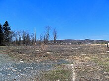 A bare expanse of untended grass and shrubbery with some bits of stone embedded in the ground. At left is a patch of gravel and at the bottom center the remnant of a curb. In the background, at center left, are some bare trees, evergreens, and a telephone pole