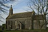 Small stone building with arched doorway. The church is partially obscured by trees.