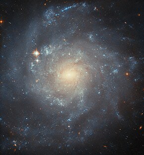 A spiral galaxy, seen up close and face-on. It is coloured yellow and glowing brightly at the oval-shaped centre, showing older and cooler stars, and it becomes bluer out to the edge of the disc where the stars are younger and hotter. It has a number of somewhat patchy spiral arms curling around, with sparkling areas where stars form. The black background can just be seen at the corners.