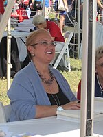 A woman with short blond hair, wearing a blue sewater and a black shirt also wearing a necklace