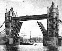 A ship with two tall masts is passing beneath the raised carriageways of a road bridge. The bridge has twin ornamental stone towers which are connected by a walkway high above the river.