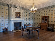 A sitting room decorated with panels painted with a 3D effect, containing a few pieces of furniture and a portrait by Thomas Gainsborough hanging over the fireplace.