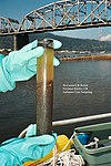 A diver’s blue-gloved hands holds a cylindrical sediment core. The core is inside a plastic tube through which dark sediments and murky water can be seen. Image text reads “McCormick & Baxter Portland Harbor, OR Sediment Core Sampling”. In the background there is the deck of a boat, water, a bridge, and a forested ridge.