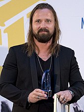 A Caucasian man with long hair is posing and smiling